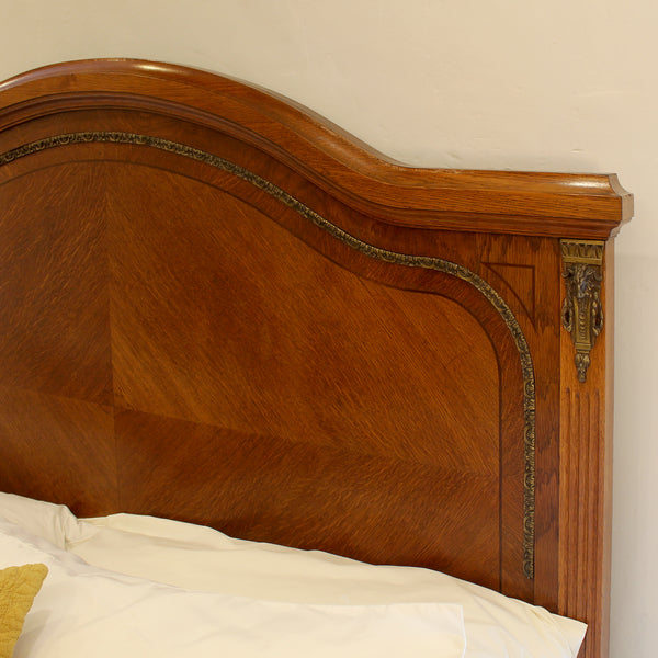 Close-up of a polished wooden bed headboard with curved detailing and brass hardware, shown above white bedding against a plain wall.