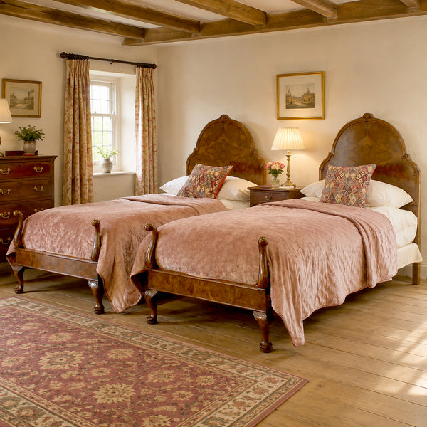 A rustic bedroom featuring two twin-size wooden bed frames with carved heads and tails, dressed in dusty pink velvet bedding and patterned pillows, set against a warm cream wall with exposed wooden ceiling beams.
