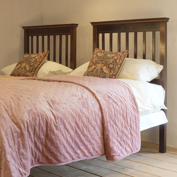 A twin bed with a dark brown wooden headboard and footboard, dressed in white bedding and a dusty pink quilted blanket, with two patterned accent pillows against a neutral beige wall.