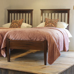 Two wooden twin beds with dark brown slatted headboards and footboards, dressed in dusty pink blankets and white pillows, with patterned accent pillows, set on a rustic wooden platform against a plain wall.