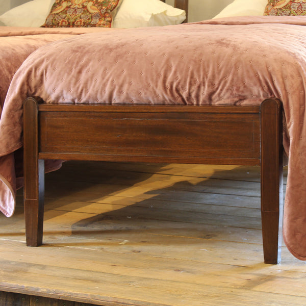 A dark brown wooden bed frame with a footboard, shown in a bedroom setting with a dusty pink velvet comforter and wooden floor.