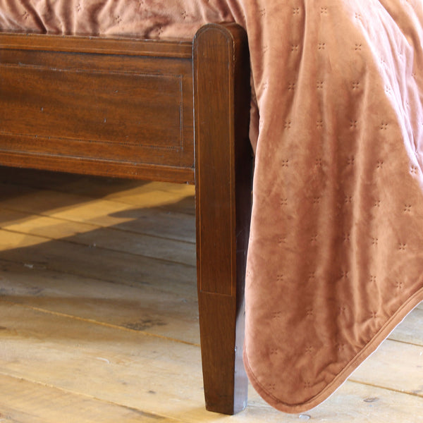 Close-up of a wooden bed frame corner with a dusty rose velvet quilt draped over the side, set on a light wood plank floor.