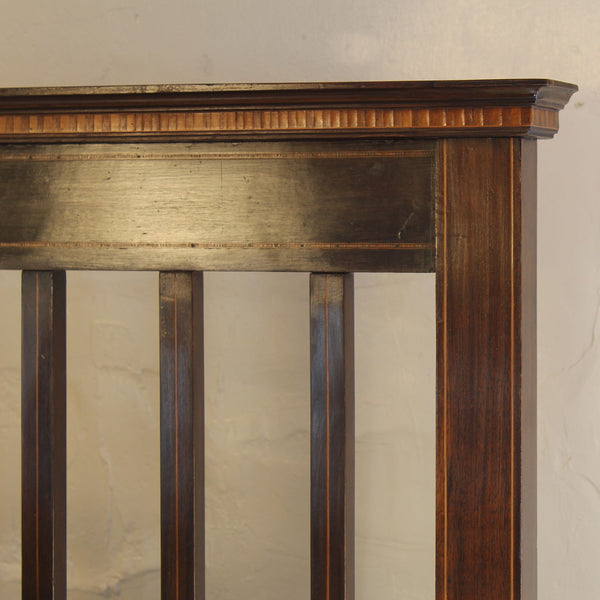 Close-up of a dark brown wooden furniture piece, showing the top rail with a decorative reeded pattern, vertical slats, and a solid front panel against a light textured wall.