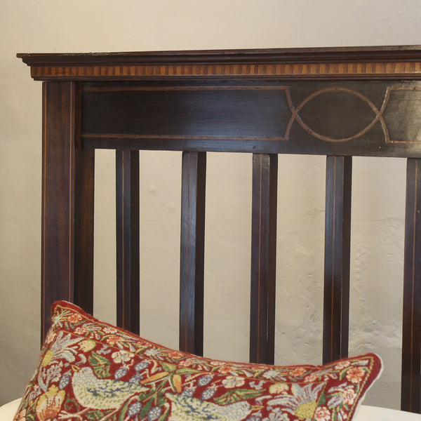 Close-up of a dark brown wooden bed headboard with vertical slats and decorative carved details, with a red patterned throw pillow partially visible in the foreground against a light neutral wall.
