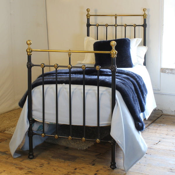 A vintage-style metal bed frame with black-painted bars and polished brass/gold accents, shown made with white bedding and a dark navy throw blanket in a rustic room with wooden floors.