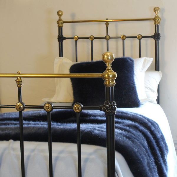 A close-up of a bed with a black metal frame featuring ornate gold-toned accents, dressed in white bedding and a dark navy blue faux fur throw and pillow.