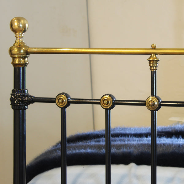 Close-up of a black metal bed frame with ornate brass/gold accents, including a round finial on the headboard post, decorative brackets, and round joint caps, with a dark blue blanket partially visible underneath.