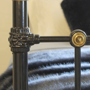 Close-up of a dark metal bed frame corner with ornate decorative brackets and brass hardware, set against a soft-focus background with a gray blanket.