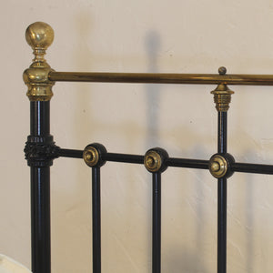 Close-up of a black metal bed frame with ornate brass accents, including a round finial on the post, decorative brackets, and round joint caps, set against a plain beige wall.