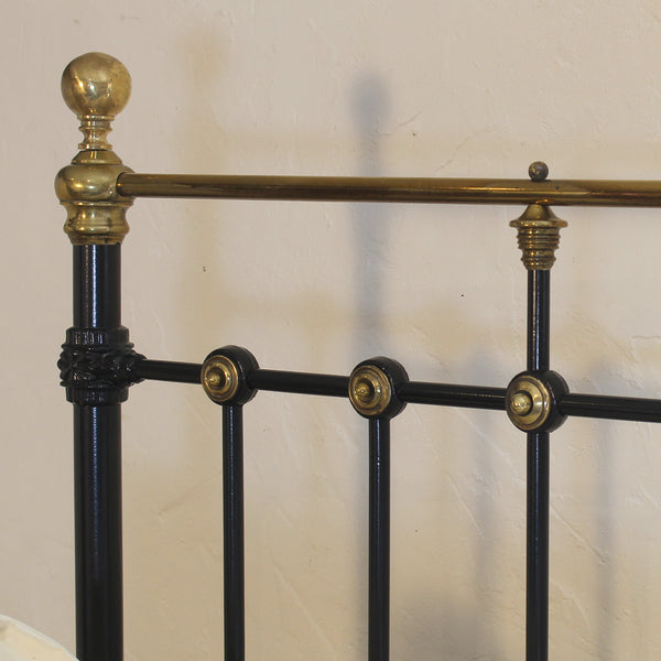Close-up of a black metal bed frame with ornate brass accents, including a round finial on the post, decorative brackets, and round joint caps, set against a plain beige wall.